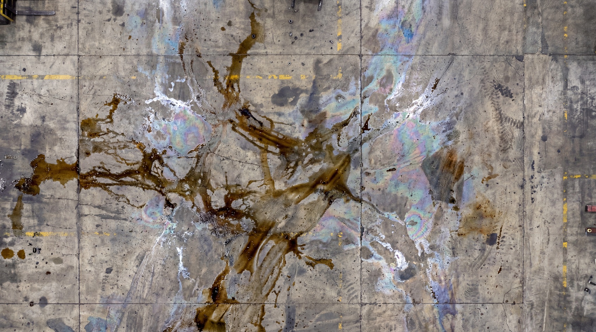 Overhead view of a concrete industrial floor with dark oil spills and iridescent rainbow patterns.