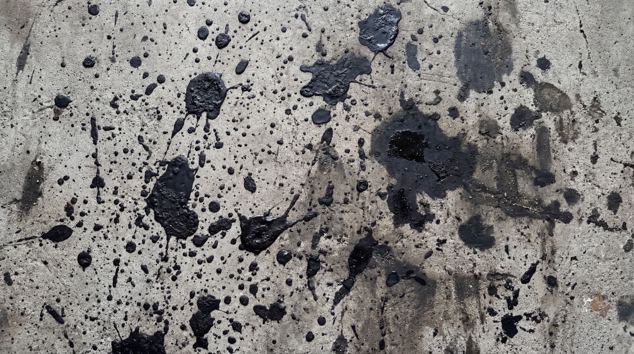 A close-up, top-down view of a raw concrete surface covered in numerous black oil spills and splatters.