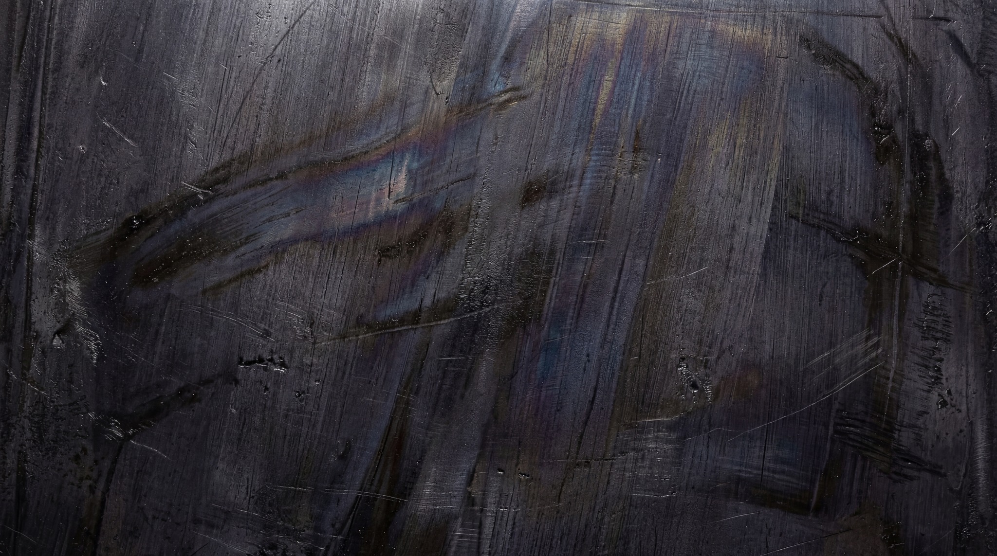 Close-up of a dark, industrial steel surface featuring heavy brush strokes, fine linear scratches, and deep metallic charcoal tones.