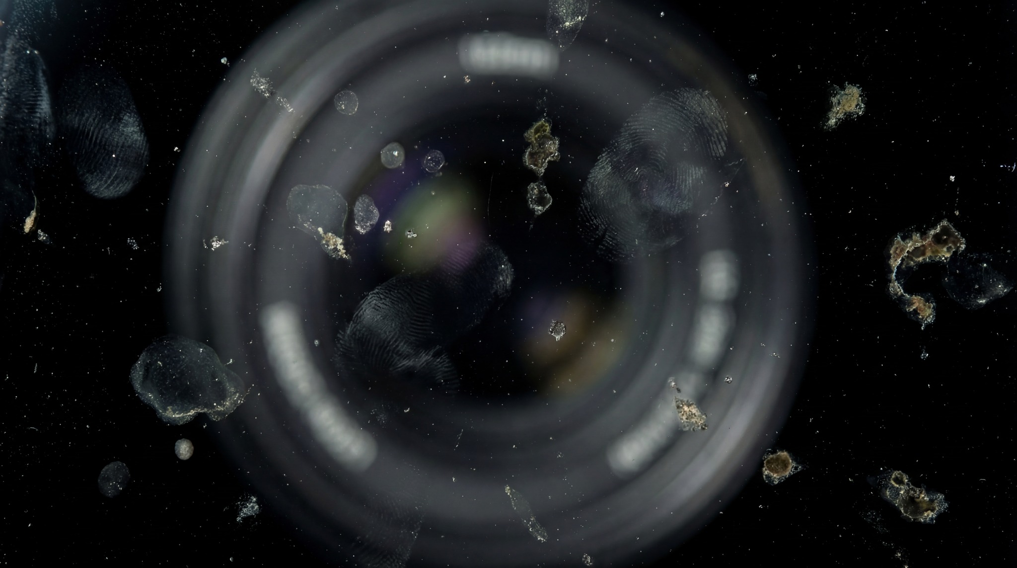 Macro shot of a dirty camera lens with visible fingerprints, smudges, and debris against a black background.