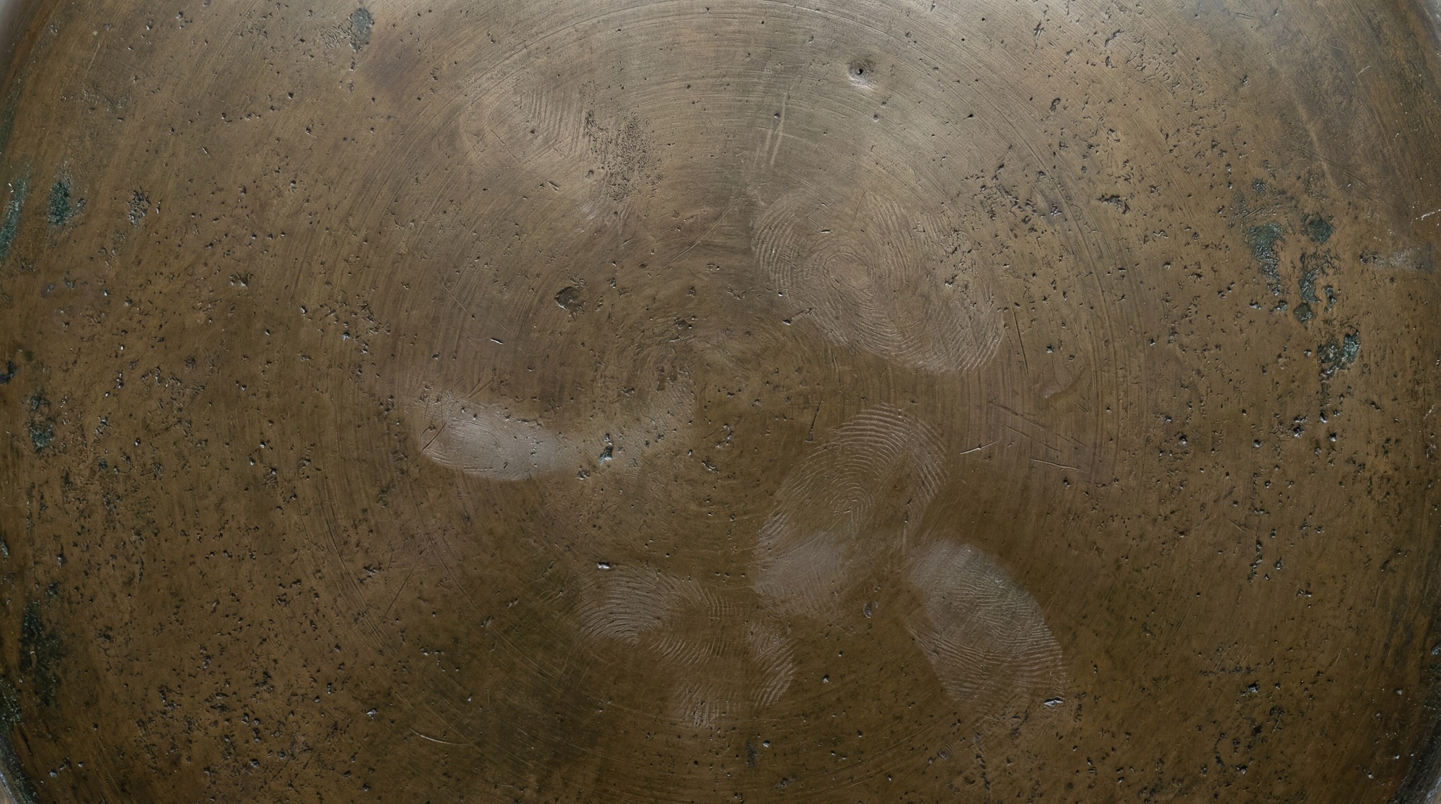 A close-up view of a weathered bronze surface featuring a dark brown patina, subtle circular scratch patterns, and visible oil-based fingerprint residue.