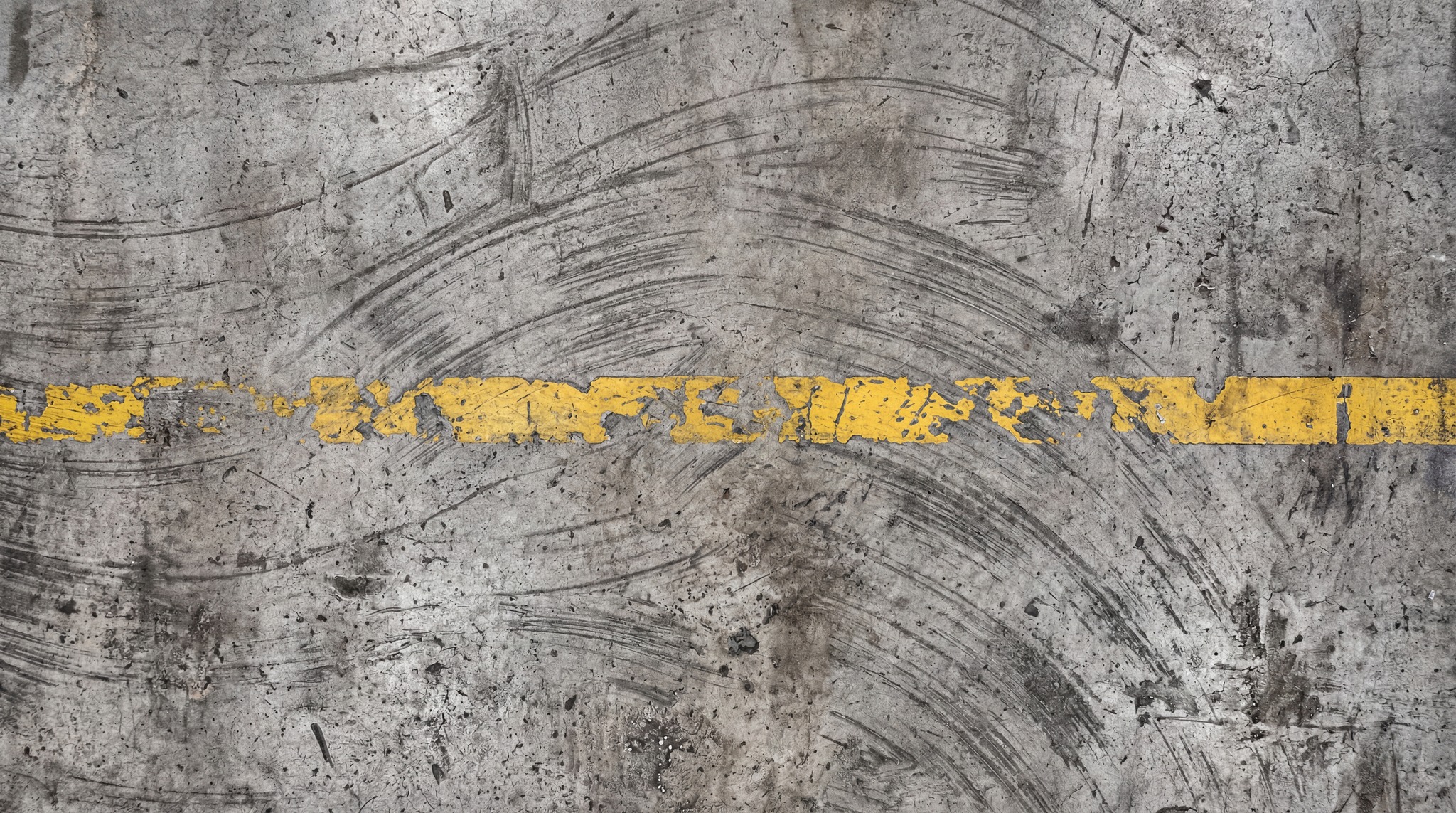 A close-up, top-down view of a textured industrial concrete floor with a broken yellow line painted across it. The concrete surface features numerous scuff marks, tire tracks, and some debris, suggesting heavy use.