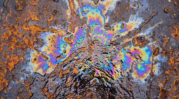 Close-up of a dark, heavily rusted metal surface with a vibrant, iridescent oil slick pattern in the center.