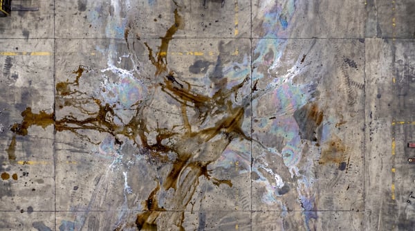 Overhead view of a concrete industrial floor with dark oil spills and iridescent rainbow patterns.