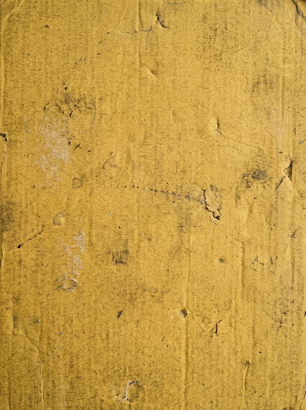 Close-up of a yellow painted cardboard surface with visible texture, scratches, and dark smudges.