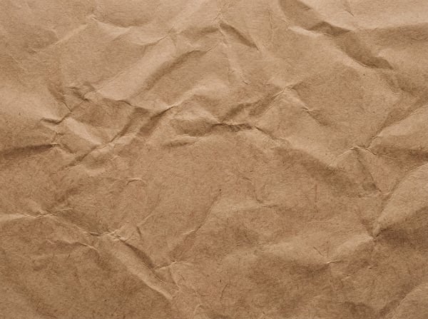 A close-up, top-down view of a crumpled sheet of light brown kraft paper, showing prominent wrinkles and folds.