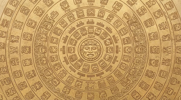 Close-up of an ornate, circular Mayan calendar carved into a textured gold surface, featuring glyphs and numerical elements.