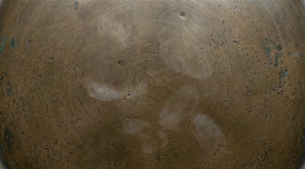 A close-up view of a weathered bronze surface featuring a dark brown patina, subtle circular scratch patterns, and visible oil-based fingerprint residue.