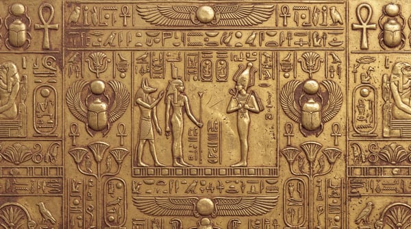 Close up of a metallic gold surface embossed with ancient Egyptian deities and hieroglyphics in a bas-relief style.