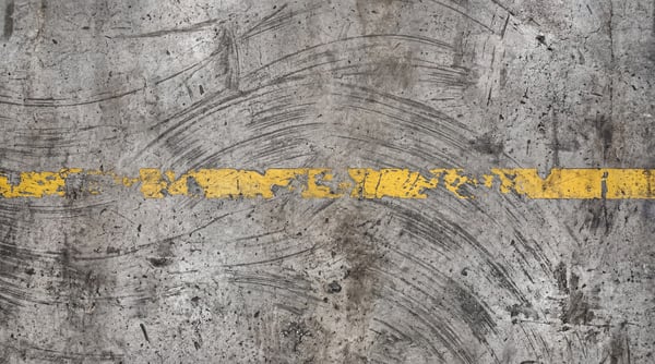 A close-up, top-down view of a textured industrial concrete floor with a broken yellow line painted across it. The concrete surface features numerous scuff marks, tire tracks, and some debris, suggesting heavy use.